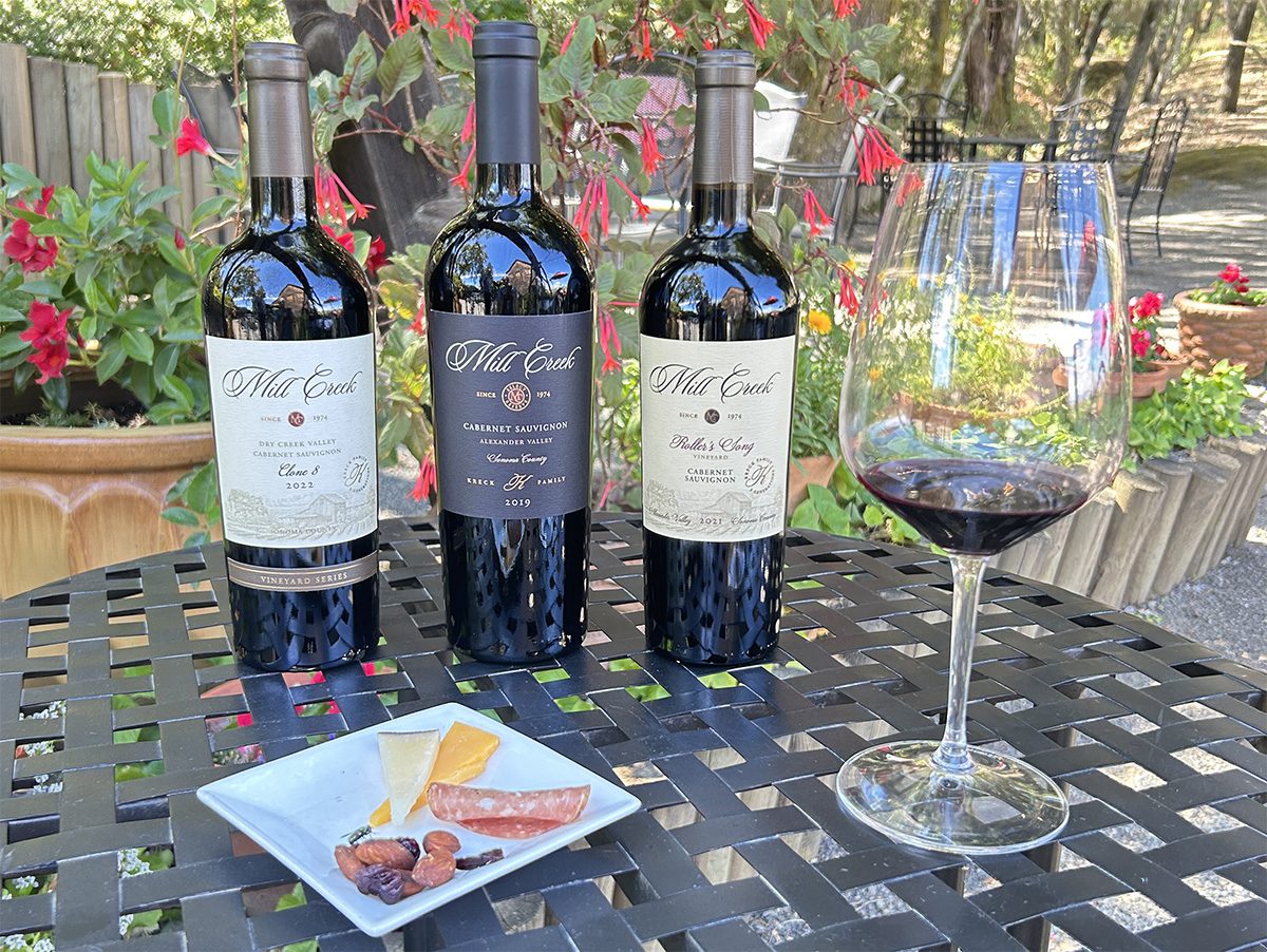 Three Mill Creek Cabernet Sauvignon bottles from our Dry Creek Valley and Alexander Valley vineyards on a patio table with a glass of red wine and a plate of cheese, salami and olives